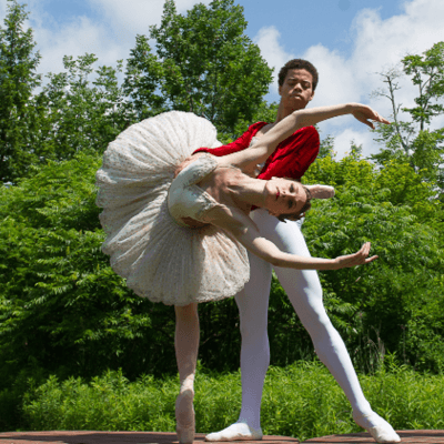Two ballet dancers pose outdoors with trees visible behind them. The female dancer on the left wears white and leans her torso and head towards the camera, while the male dancer wearing red on her right supports her.