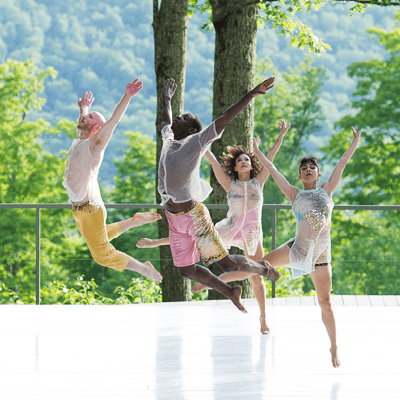 Four dancers performing in an open air venue with trees behind them.