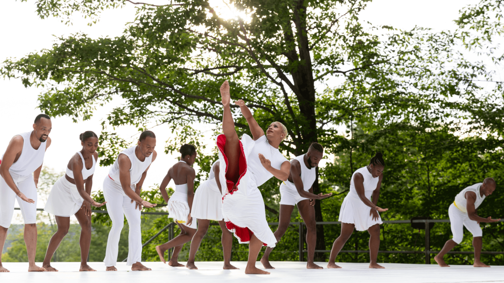 A dancer wearing a white dress with red lining raises their right leg to the sky with seven dancers in white behind them on an outdoor stage. Trees are visible behind them.