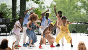 A group of dancers wearing loose fitting, bright colors poses in the center of an outdoor stage. Audience members are visible in the foreground.