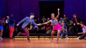 Nathan Bugh and LaTasha Barnes in motion grooving excitedly with a hand connected to each other. Behind them is a large band with dancers around cheering them on. Photo by Grace Kathryn Landefeld.