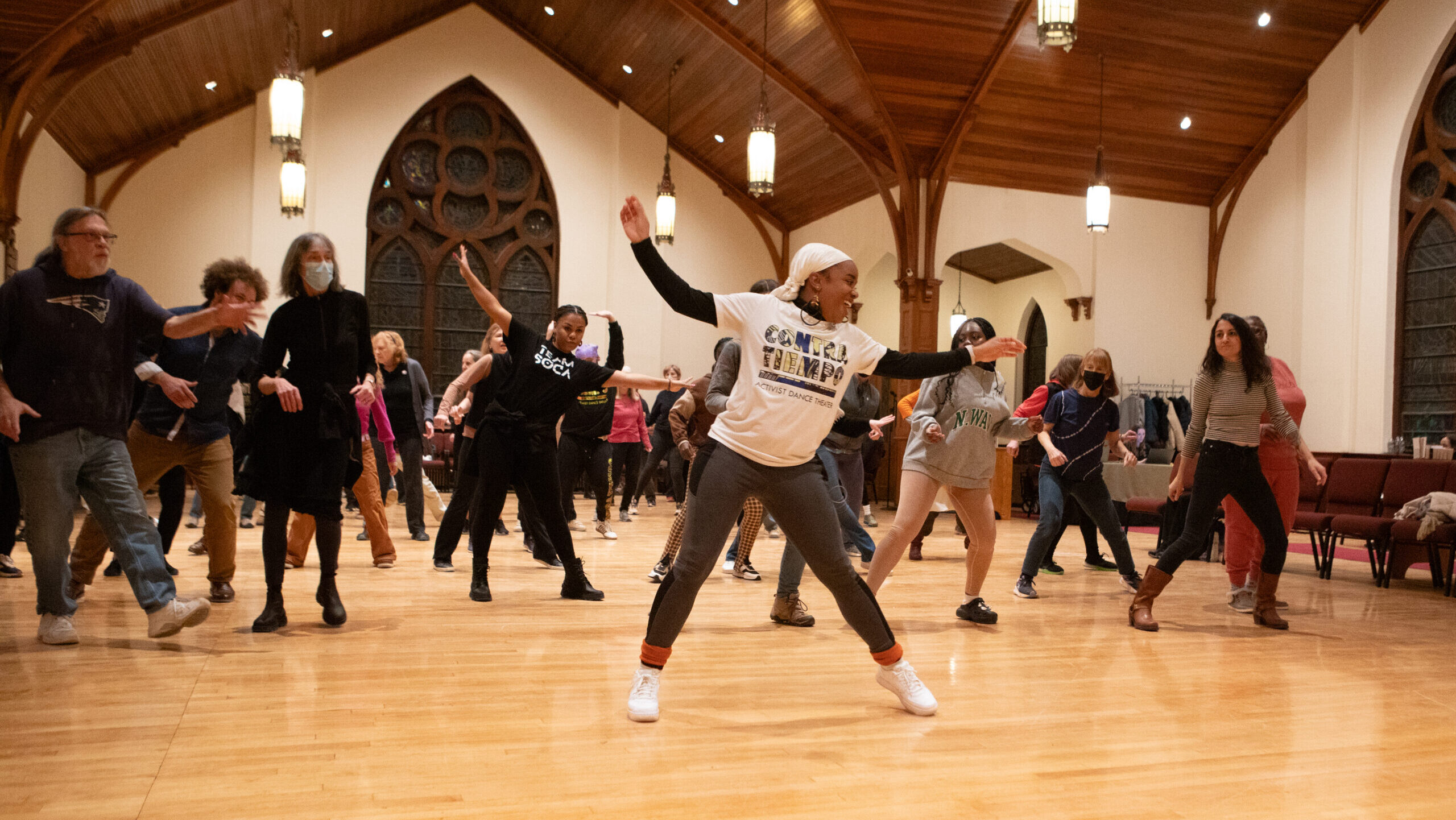 Participants of diverse ages and backgrounds are actively engaged in a dance class in a large Church dance studio with arched windows and wooden floors. The dance instructor stands in the forefront wearing a white headscarf, white Contra-Tiempo t-shirt, and gray pants while striking a pose.