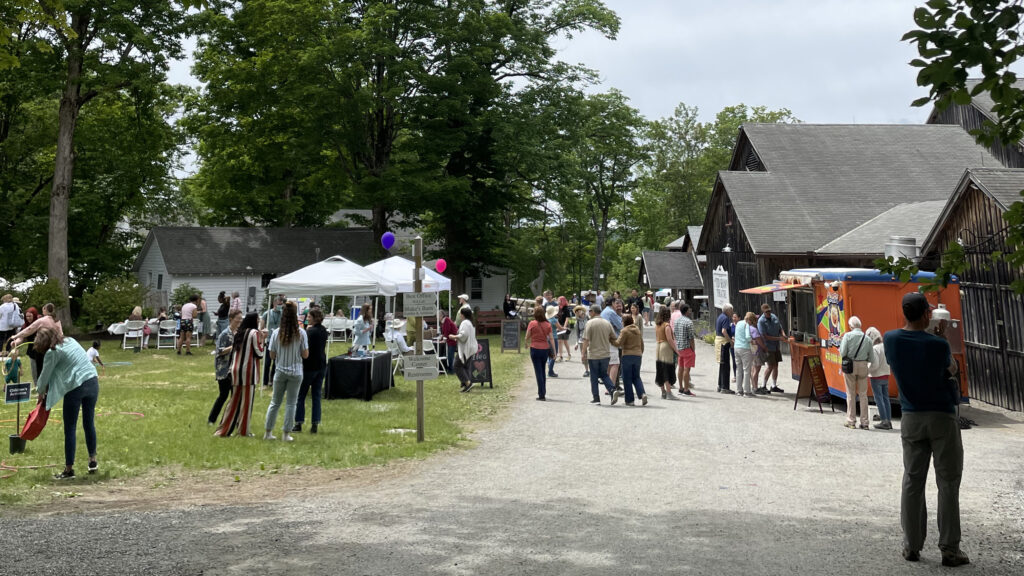 Patrons stroll on the gravel main path between a wooden theater and a large grassy lawn.