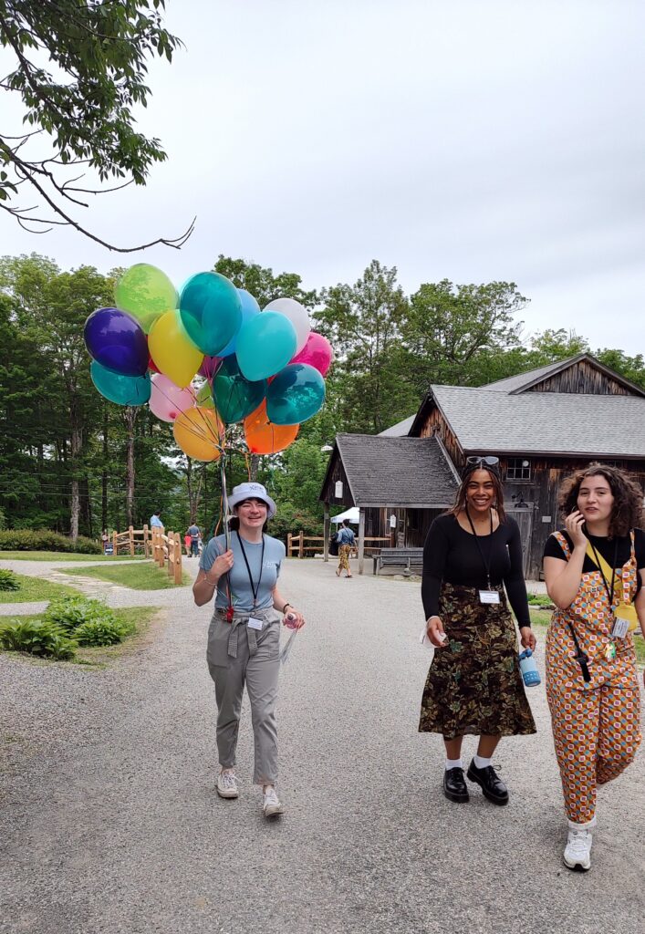 Three people walk up a gravel path. One wearing a blue shirt carries a bunch of colored balloons.