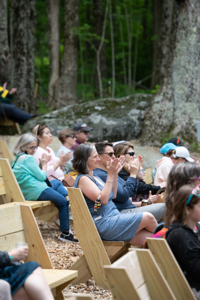 Audience members sit and clap in outdoor benches.