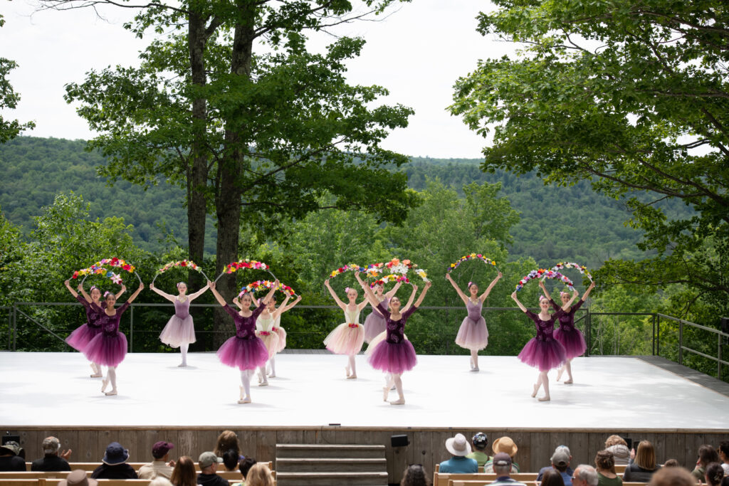 A group of dancers wearing romantic tutus in varying shades of pink and purple face the audience on an outdoor stage. They cross one leg behind the other, as if about to curtsy, and hold flower garlands above their heads.