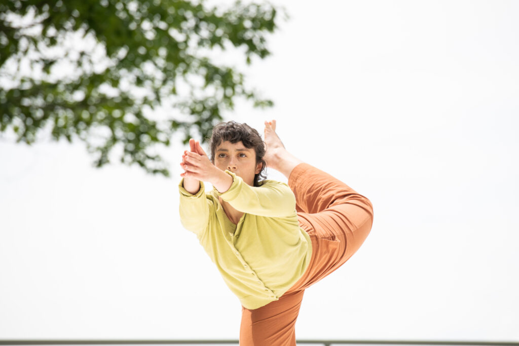 A dancer wearing a lime green sweater and orange pants stands on her right leg and shoots her arms forward. Behind her, her left leg curls up into the air.