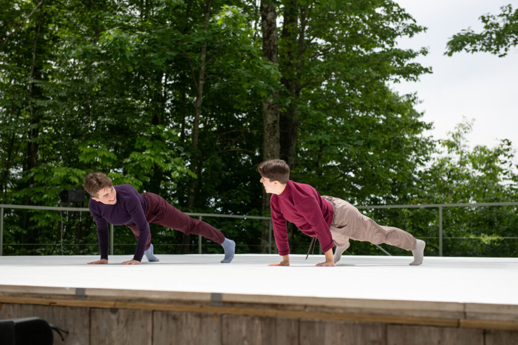 Two dancers are in a plank position on an outdoor stage. They are wearing colorful turtlenecks and looking at each other smiling.