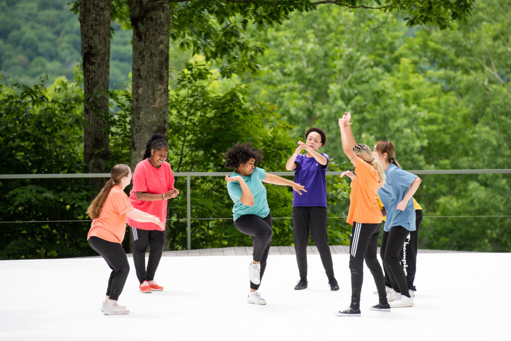 A group of dancers on an outdoor stage wear colored tshirts and black pants. They are facing each other and in various poses of joy. 