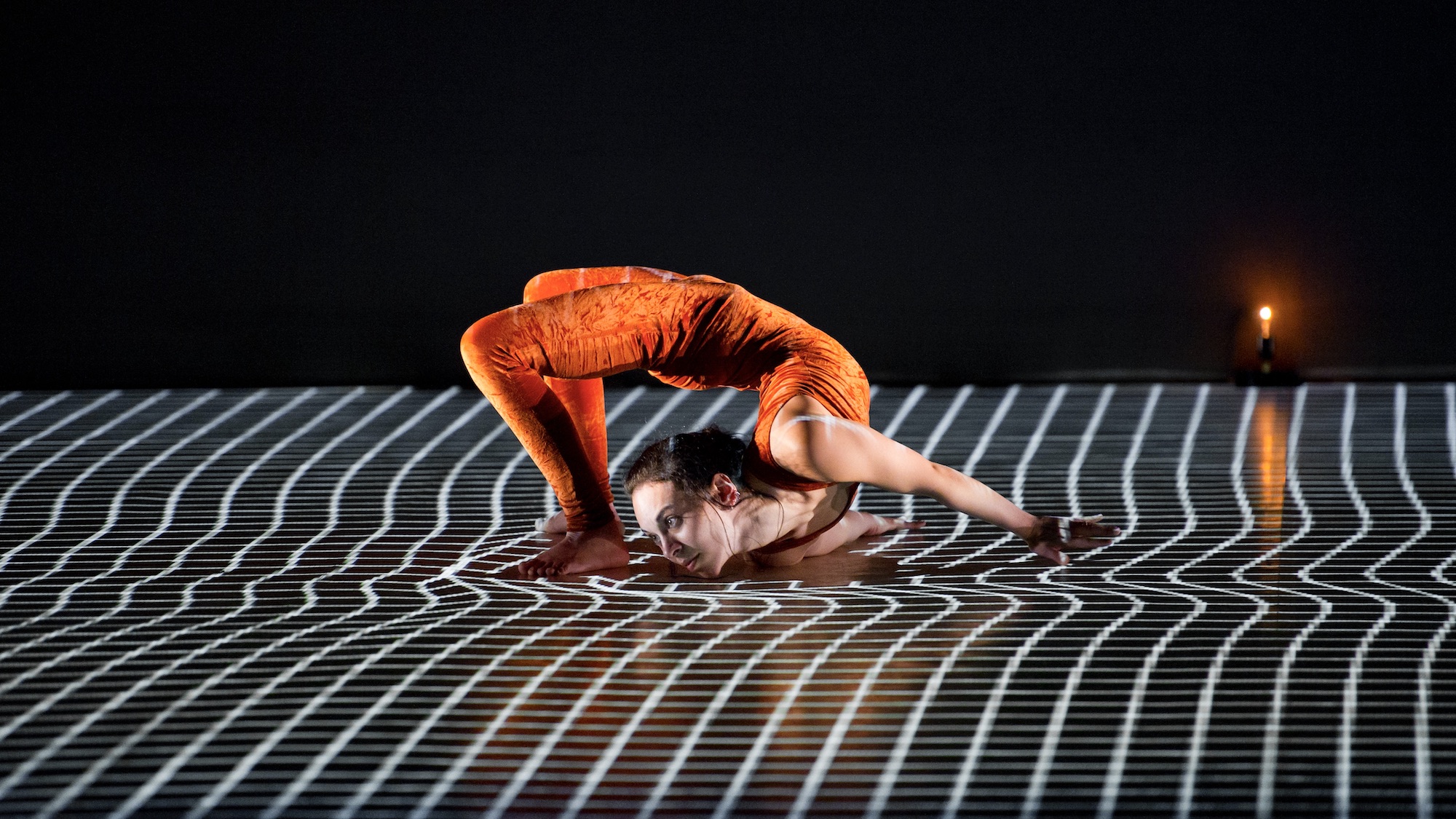 A dancer in an orange unitard contorts her body on a floor with white twisting lines on it.