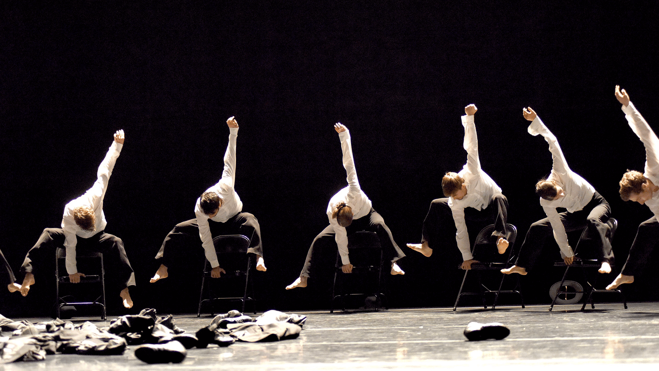 Dancers of Gauthier Dance//Dance Company Theaterhaus Stuttgart photographed in motion holding themselves in the air with one hand on top of black chairs.