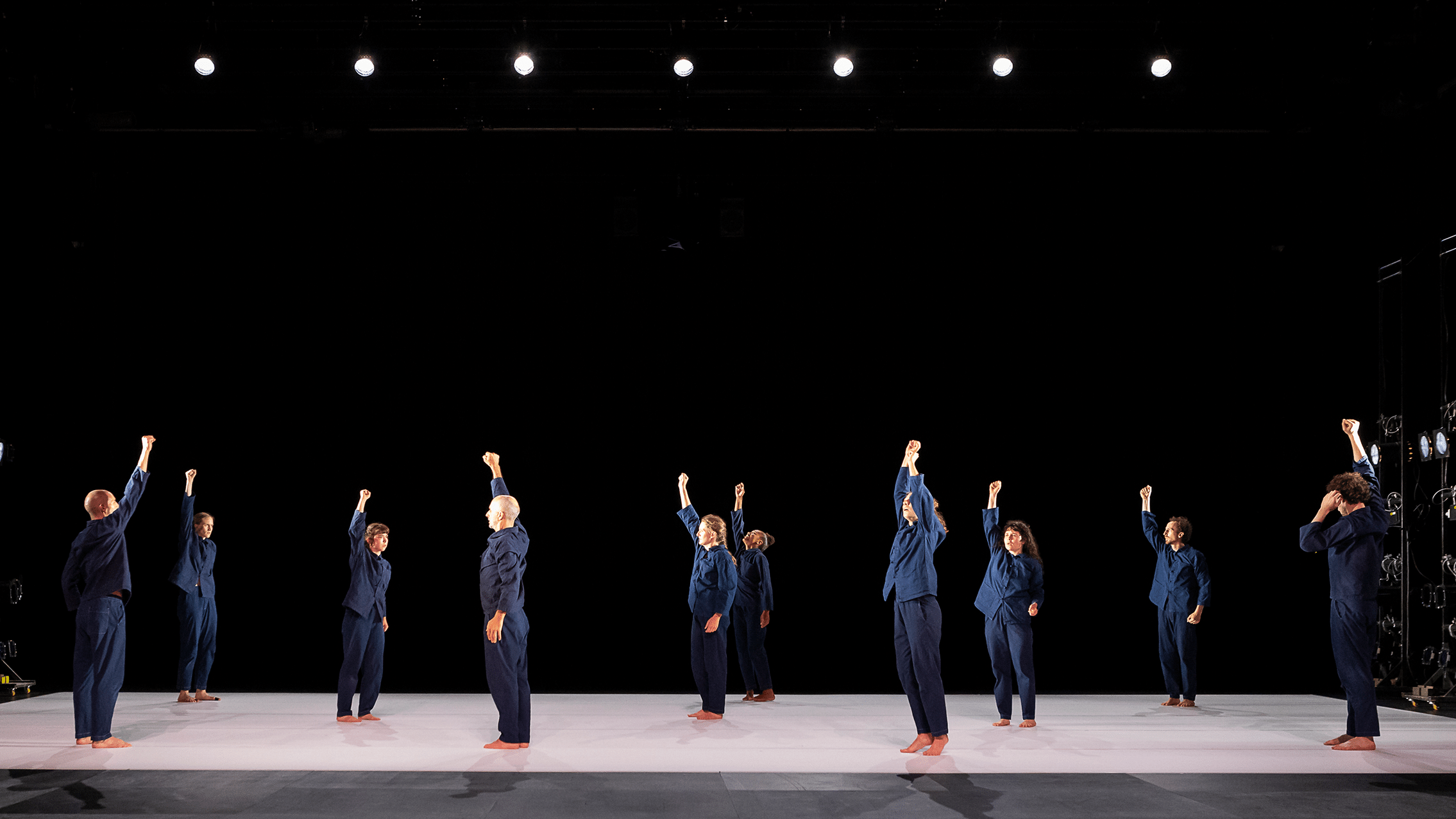 Dancers of Oona Doherty scattered across a stage facing different directions with their right arm extended to the air.