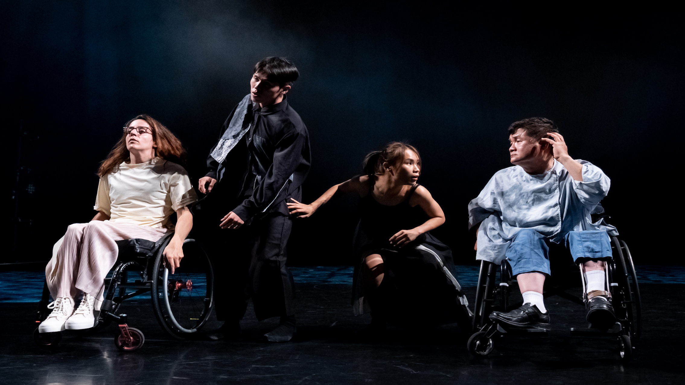 A woman in a wheelchair, a standing man, a crouching woman, and a man in a wheelchair all pose on a stage.