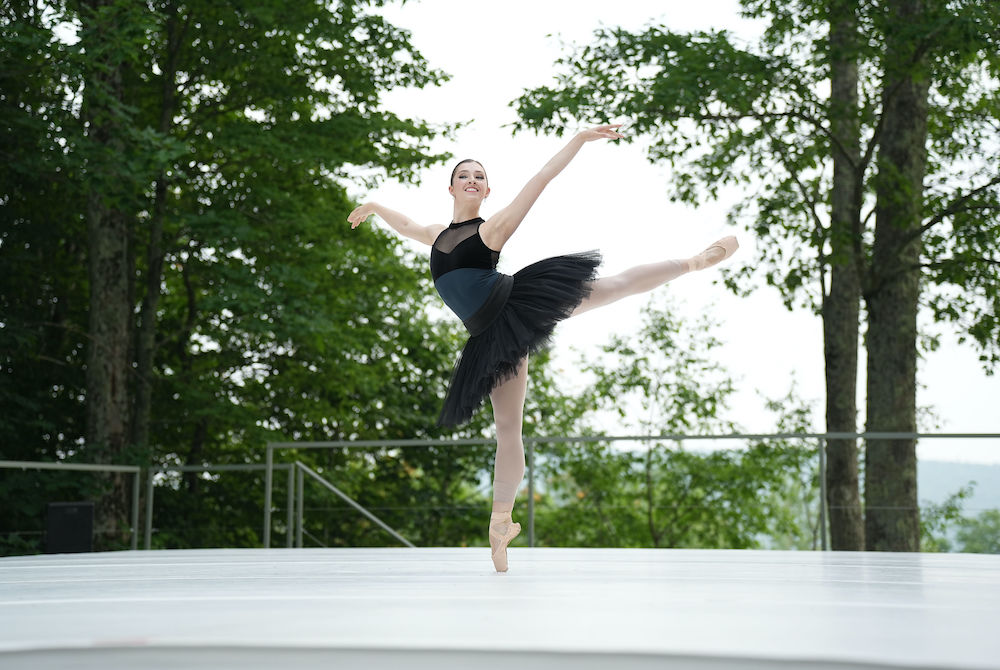 A dancer wearing a black tutu stands on pointe and holds her arms out.