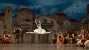 One dancer in a white costume leaping center stage while 11 additional dancers look on.