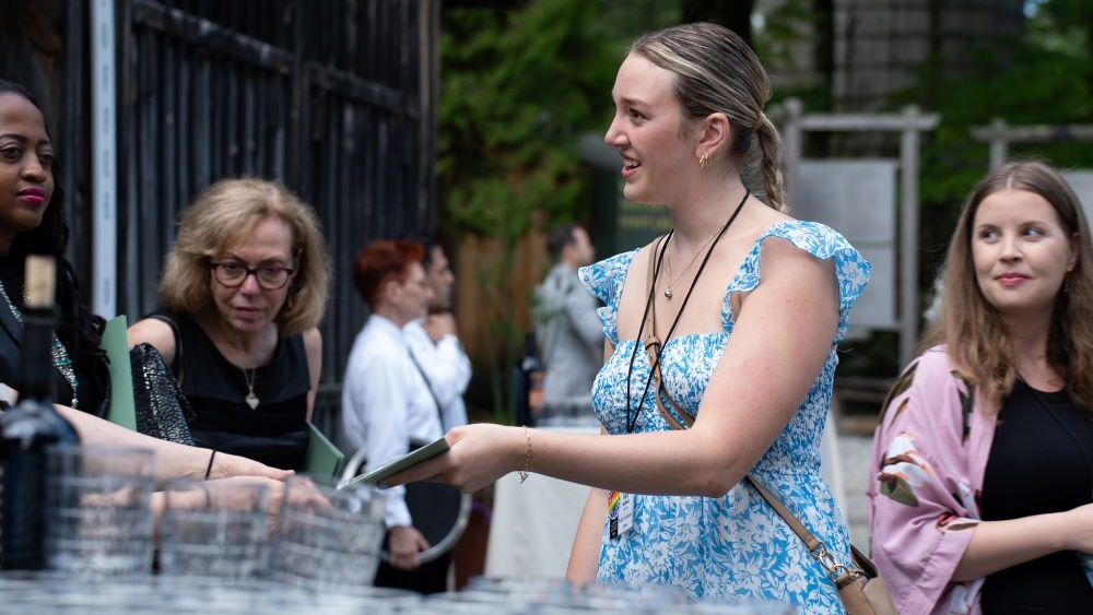 A young, blonde woman wearing a blue floral dress is pictured handing a document to a person off camera.