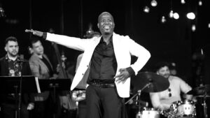 Sekou McMiller, a bald, Black man, appears in black and white on a stage with musicians behind him. He is wearing a white suit jacket, a black button down, and black pants.