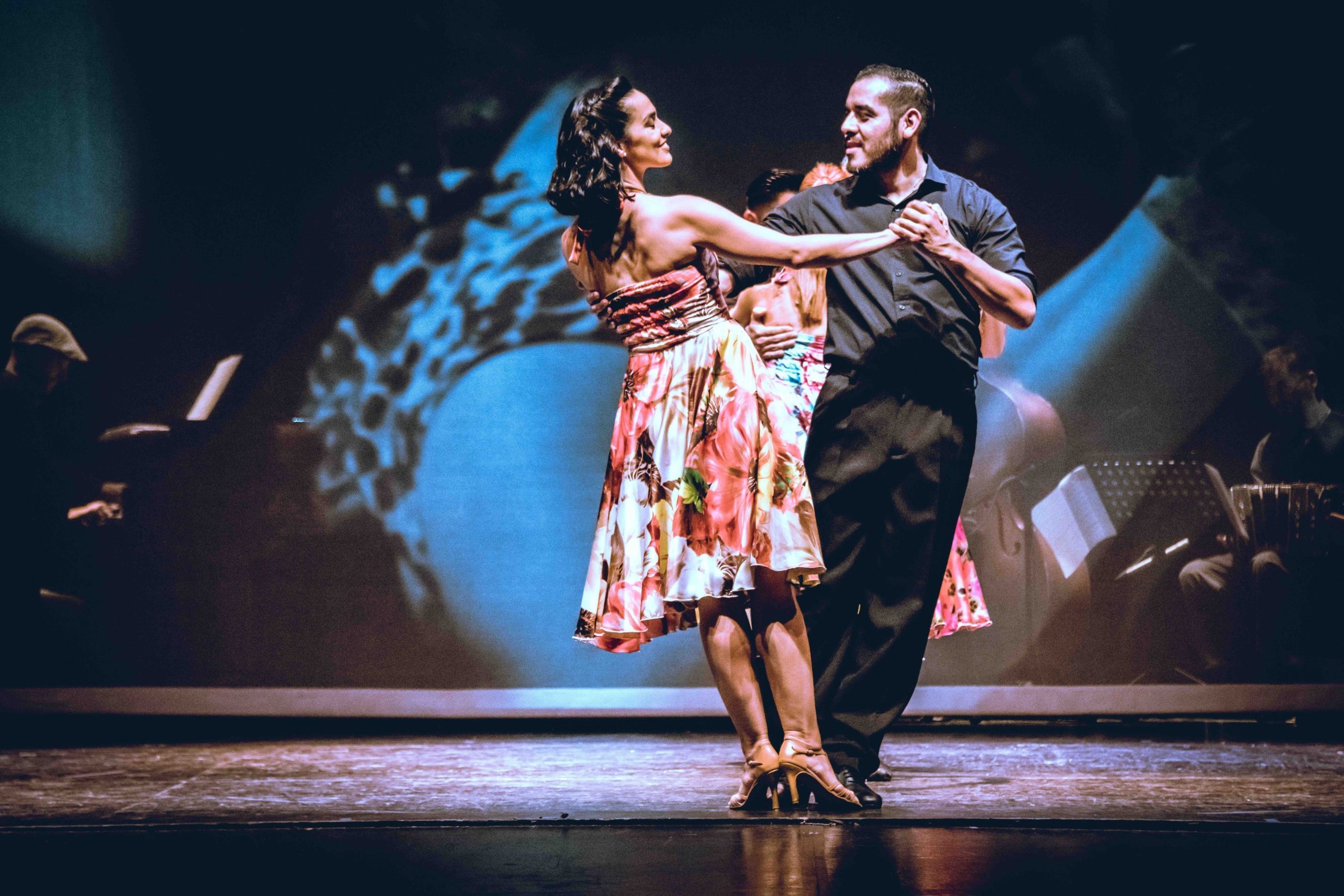 A pair of tango dancers appear on a dimly lit stage. He is wearing dark pants and a button-down shirt, and she is wearing a floral dress. They are dancing in each other's arms with smiles on their faces.