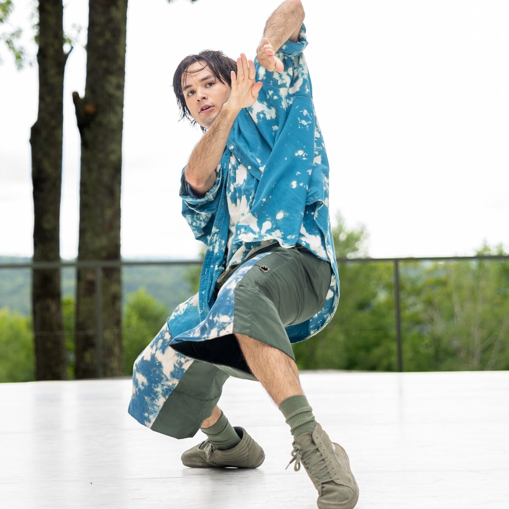 A dancer wears a blue, baggy shirt with light bleach spots and green shorts with the same blue fabric on front. The dancer balances on the inside of one foot and the toe of the other foot while their arms are bent close to their face and their fingertips point away from them, like blades. If they extended their arms, it would look like they were about to swim the breaststroke.