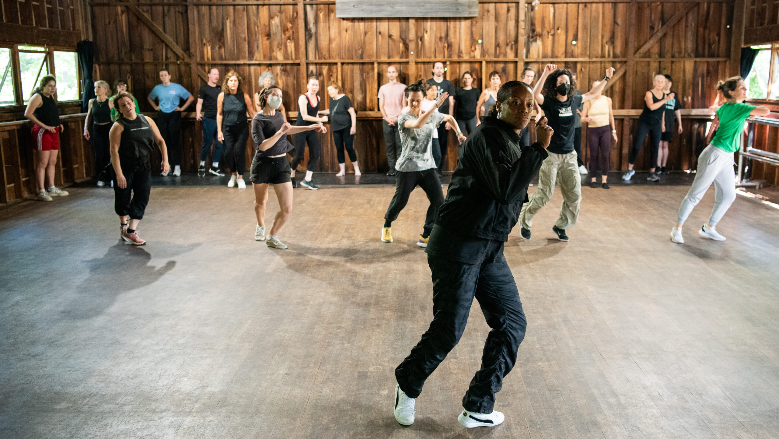 Inside a wooden studio, a teaching artist wearing all black demonstrates a dance move. Five students are in a line behind the teacher while mimicking the dance move, and the remaining students are leaning against the wall and watching.
