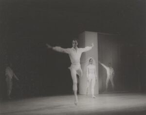 Merce Cunningham, dressed in white tights and a white billowing shirt, jumps in the air with one leg extended back and arms out in a "T." Behind him are three other dancers in white. One is a woman who wears a form-fitting white leotard with long sleeve and turtleneck, and light colored tights. On her head is a fluffy, veil made of tulle.