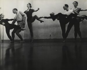 Six dancers pose, creating two groups. There are three people in each group creating a partnered shape by layering their bodies in front of and behind each other. They are dressed in casual clothing.