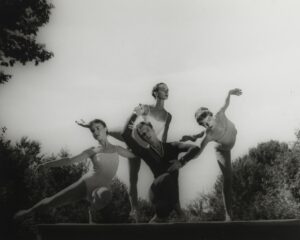 Four dancers pose on an outdoor stage-like platform. Merce Cunningham, dressed in a dark full-length, long-sleeve unitard, crouches low with legs crossed. One arm is bent above his head, the other bent in front of his body. Three women dancers use his arms for support. They are dressed in thin-strap light-colored leotards. One woman stand behind him, balancing on one leg. The others stand on either side of him, one balancing on a crouched leg, the other balancing on a straight leg while leaning towards Cunningham.