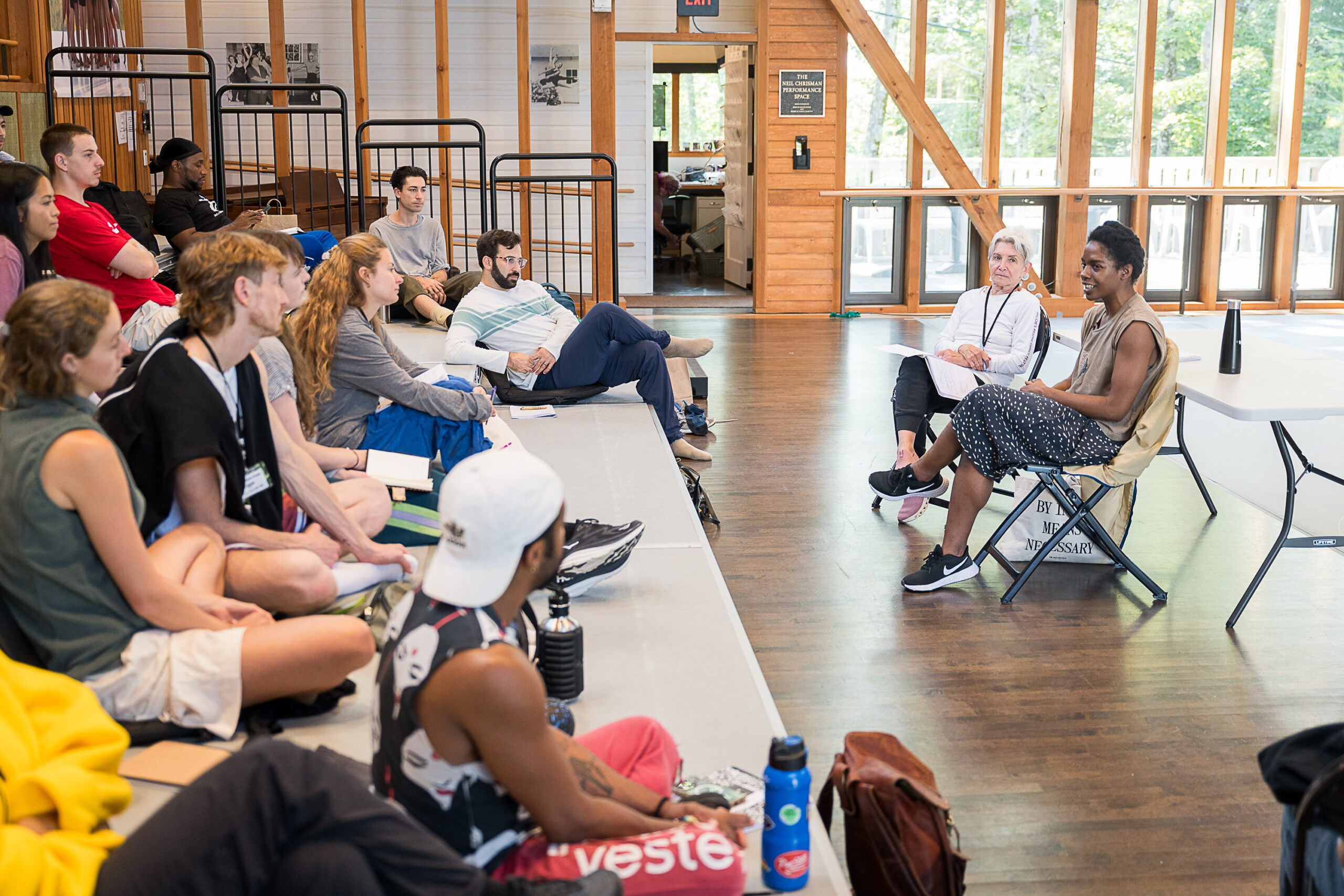 In a wooden studio, two people sit in folding chairs, facing a group of dancers sitting in the rises in front of them. The two individuals are speaking to the group, while the group listens intently.