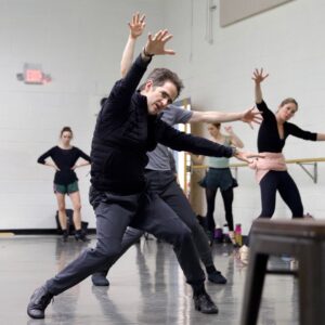 A dancer in a studio reaches his arms out in a "V" around his head, with his legs bent. 