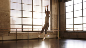 Emily Coates, dressed in black leggings with a white stripe on the side of the leg, a long sleeved gray shirt, and white crew socks, leaps into the air with her arms high above her body, reaching to the sky and her legs and feet pointed downward. She is dancing in a large room with marley on the floor and giant, luminous windows where the sun is glaring in.