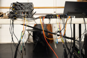 A desk with several multi-colored wires streaming out of several computer systems, inside of a dance studio.