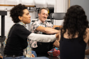 A man with gray hair and a collared shirt sits on the floor, smiling in conversation at two out of focus people in the foreground.