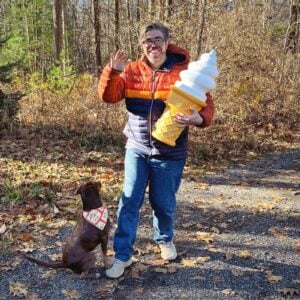 Eryk, a white man with brown hair, beard, and glasses, holds a large plastic sculpture of a soft-serve ice cream cone in the woods with a small brown dog.