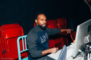 André, a Black man with black hair and beard sitting on a blue-painted metal chair in front of a computer pointing his left hand and finger to the side wearing a grey & blue zip up sweater.