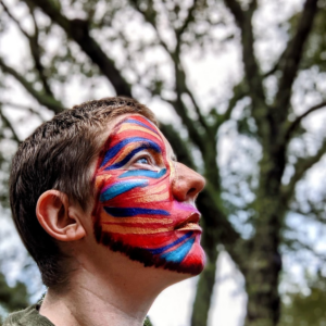 Laurel looks up against the branches of a massive oak. Their face is painted red with lines and curves of turquoise, azure, and gold sweeping across lines of bone and muscle.